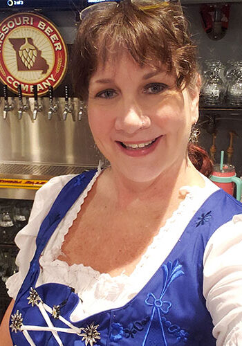Molly wearing a traditional German Dirndl, often worn by female beer servers at Octoberfest. She is standing in front of beer taps with a sign for Missouri Beer Company hanging on the wall.